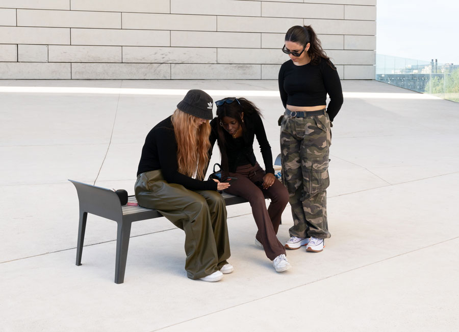 Trois jeunes femmes sur le Banc simple Ontario aluminium conçu et fabriqué par Aréa mobilier urbain